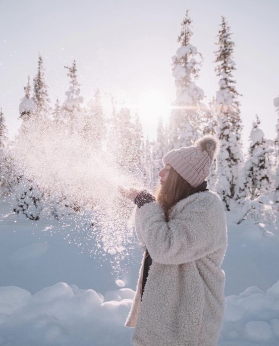 Model dressed in textured coat and beanie blowing snow outdoors with snow capped trees in the background.